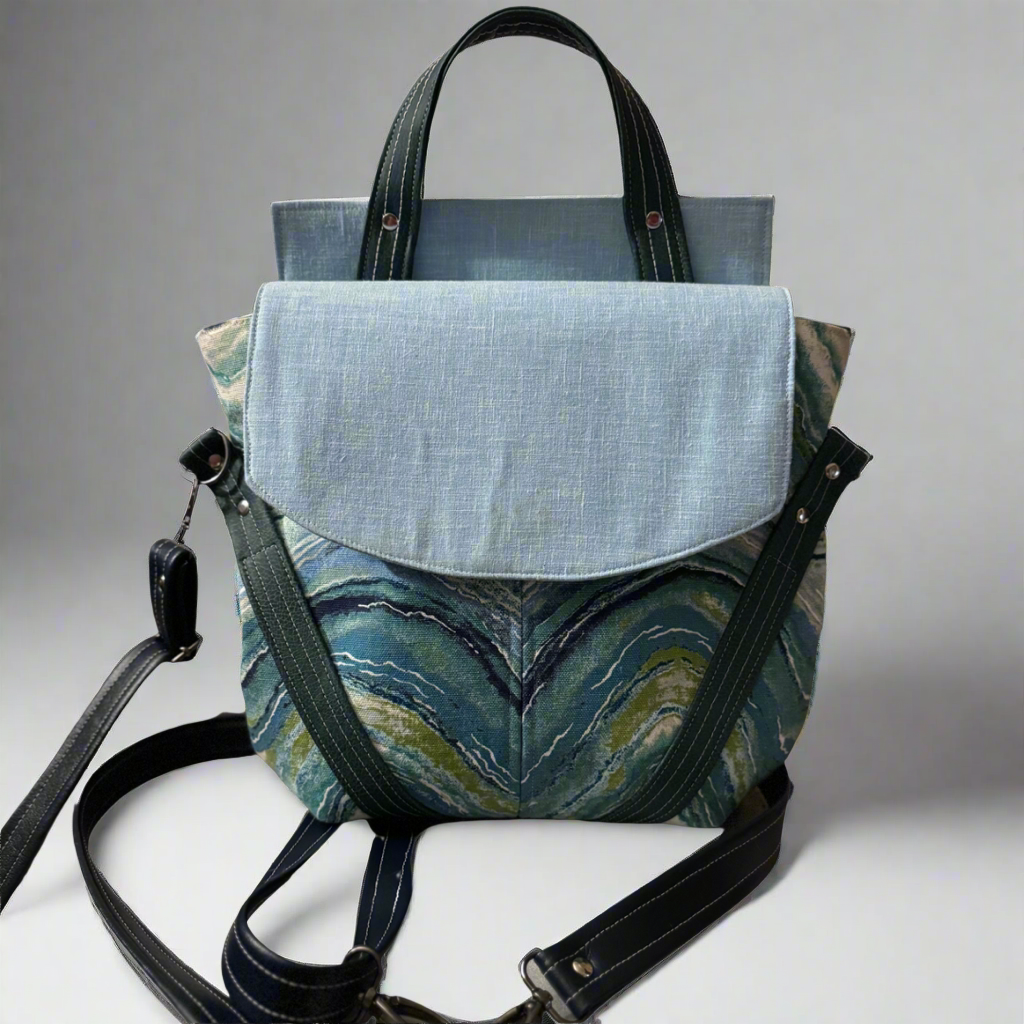 Picture of convertible handbag/backpack. It has a light blue flap, dark blue handles, strap, and accents, and a blue wave patterned front.