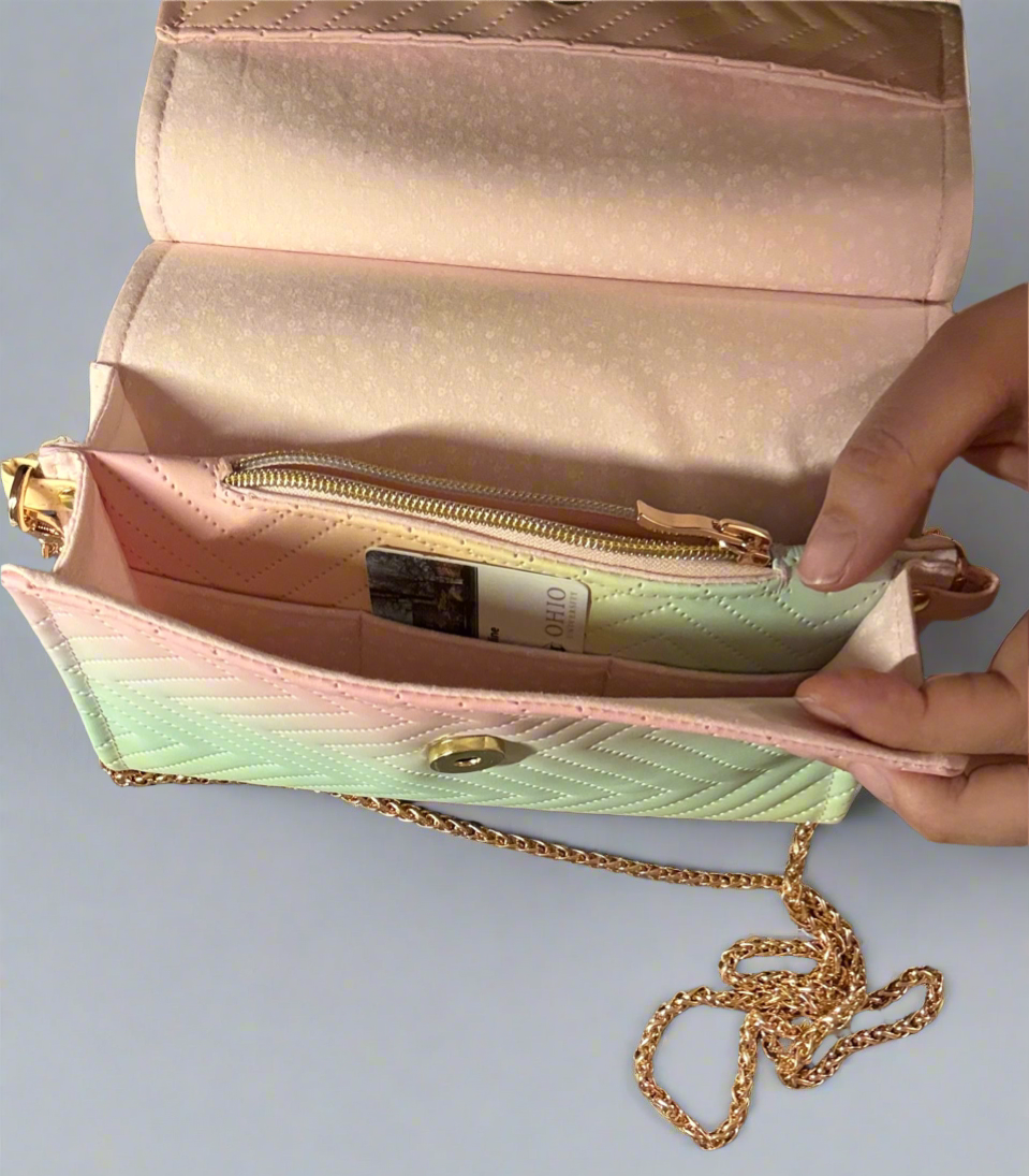 A close-up of an open, handcrafted clutch purse. The purse has a flap closure and a delicate gold chain strap. The material is quilted in a chevron design, and the colors transition smoothly from a light green at the bottom, through a pale yellow in the middle, to a very light pink on the top flap. The flap is open, revealing the inside of the purse which has a zippered compartment. There is a gold magnetic snap and a gold zipper. There are three pink card slits inside.